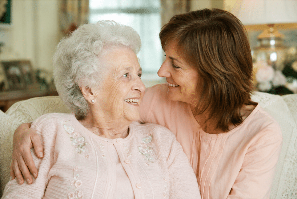 Smiling elderly woman with white hair in a pink cardigan sits on a sofa as a younger woman hugs her warmly.