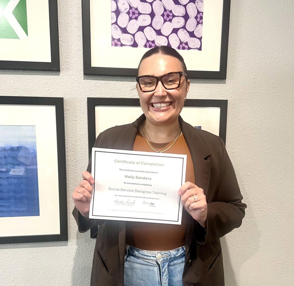 Smiling person wearing glasses and a brown jacket holds a certificate in front of framed artwork on a wall.